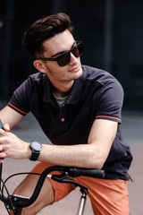 Fototapeta na wymiar Stylish guy dressed in trendy casual clothes rides bicycle in the street against the background of a gray building