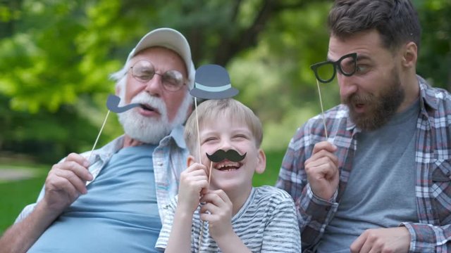 Happy funny family son and dad, granddad with fake mustache, hat, eyeglasses on holiday outdoor in park. Good day, happiness, friendship, stroll, holiday concept.