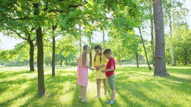 Group of three kids playing counting out game standing on green lawn surrounded by trees in park on summer day
