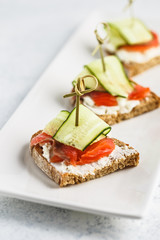 Three bruschettas with cream cheese, salmon and cucumbers on white rectangular plate on white background