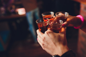 Male hands cheer with glasses of shot or liqueur. Friends drink shot or liqueur and cheers. Male hands cheer with alcohol on blurred bar background. Party and toast concept. - obrazy, fototapety, plakaty