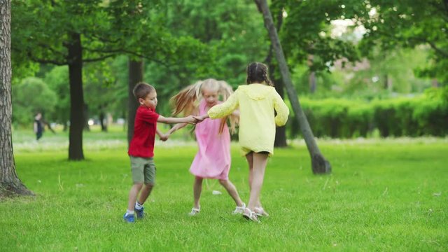 Group of three happy carefree kids round dancing holding hands on green grass in summer park