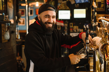 Fototapeta na wymiar smiling barman pouring beer in a bar. Bearded courageous man pours you a foaming drink. Golden tap for draft beer