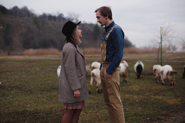 Love story. Woman in hat and her boyfriend standing next to each other at the field full of goats somewhere at countryside
