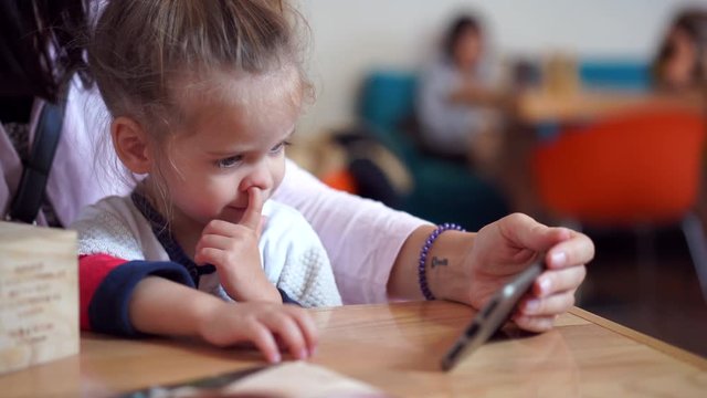 A little cute girl of European appearance is sitting on the hands of her mother at the table, watching a video on her body and picking her nose. A child with bad habits in a public place