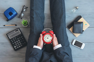 Tired overworked businessman is looking on a red alarm clock in his hands on an office floor...
