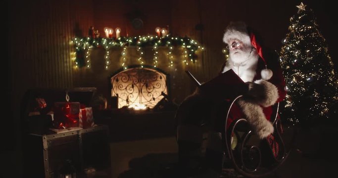 Santa claus sitting on roker in specially decorated room, reading a magical shining book - holidays and celebrations, christmas spirit concept 4k footage
