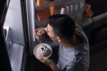 Young man eating food near refrigerator at night