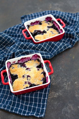 Two freshly baked blueberry cobblers in red pans on blue kitchen towel with black textured background.