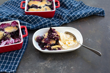 Freshly baked blueberry cobblers in red ceramic pans; some isolated on white plate in front with vanilla ice cream; blue kitchen towel; black textured bakcground.
