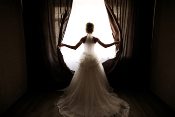 the bride in a wedding dress with a train opens the curtains of the window, stands back, you can...