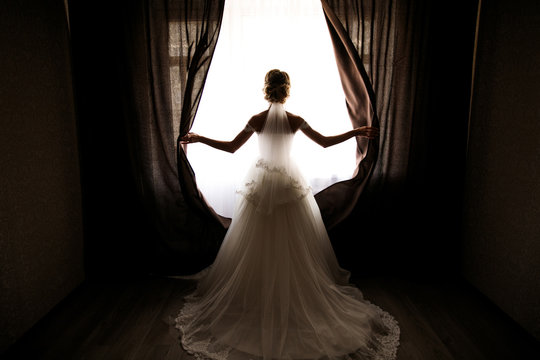 the bride in a wedding dress with a train opens the curtains of the window, stands back, you can see her silhouette, against the light