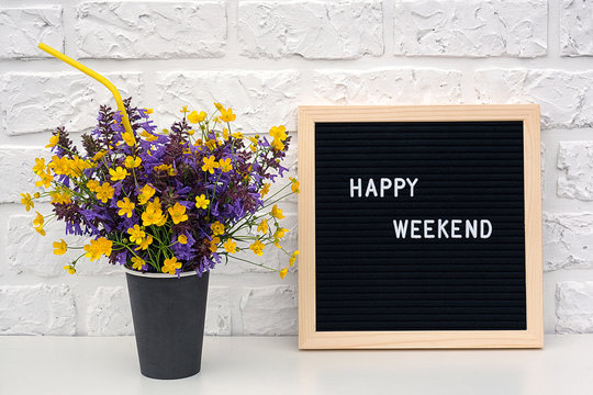 Happy weekend words on black letter board and bouquet of colored flowers in black paper coffee cup with cocktail straw on table against white brick wall. Concept Happy Weekend. Template for postcard