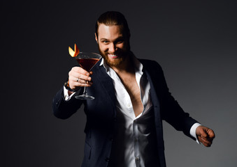 Cheerful modern bearded man in unbuttoned shirt at the party offers to try red cocktail drink with orange in martini glass