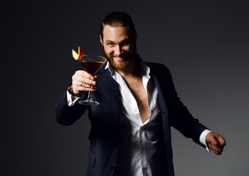 Cheerful modern bearded man in unbuttoned shirt at the party offers to try red cocktail drink with orange in martini glass