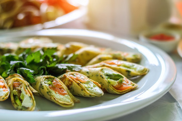 Rolls from thin pancakes with salmon and cream cheese on a white plate with fresh parsley green. Close-up, selective focus