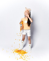 Awakened child asian girl kid in home clothes at the morning breakfast pouring out corn flakes yawning on gray 