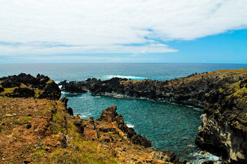 Rocky coastline near the Kai Tanata cave on Easter Island, showcasing striking black volcanic rocks, turquoise ocean waves crashing on the shore, and dramatic coastal scenery under a sunny sky.