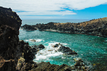 Rocky coastline near the Kai Tanata cave on Easter Island, showcasing striking black volcanic rocks, turquoise ocean waves crashing on the shore, and dramatic coastal scenery under a sunny sky.