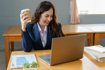Female office worker wears business suit working with laptop while drinking coffee in smiling face in the office. Business woman working and drinking coffee in relaxing manner.