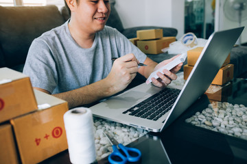 Young asian man looking on computer laptop and writing order list on paper, SME concept.