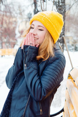 Attractive young woman with blond hair in a yellow knitting hat is warming her hands in the winter park.