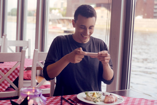 Male food blogger takes a picture on the phone from social networks. A man shooting food in a restaurant. A man makes a photo of salmon and risotto, dish. National food, tourist smile. Roaming. - Powered by Adobe