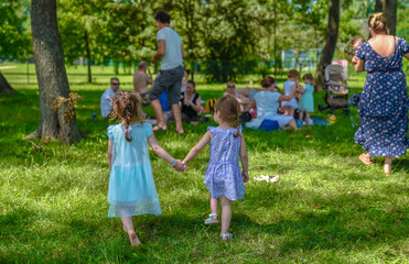 Two preschool girls in turquoise and violet dresses walking and holding hands in park while approaching a group of people having picnic at the park and enjoying nature 