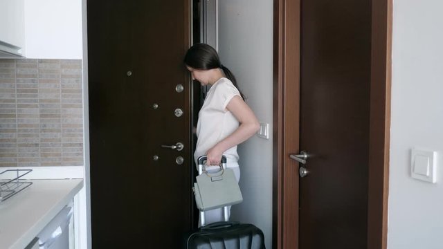 Young Brunette Woman With Suitcase Is Leaving Rented Apartments. She Goes Out The Door And Closes It Behind Her. Travel Concept.