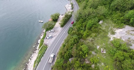 The quadcopter is flying over a narrow asphalt road running along the sea shore. We can see the turquoise sea and the seashore covered with green trees on the other side of the road