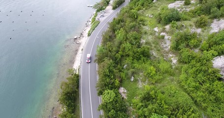 The quadcopter is flying over a narrow asphalt road running along the sea shore. We can see the turquoise sea and the seashore covered with green trees on the other side of the road