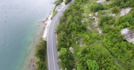 The quadcopter is flying over a narrow asphalt road running along the sea shore. We can see the turquoise sea and the seashore covered with green trees on the other side of the road