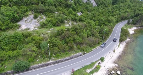 The quadcopter is flying over a narrow asphalt road running along the sea shore. We can see the turquoise sea and the seashore covered with green trees on the other side of the road