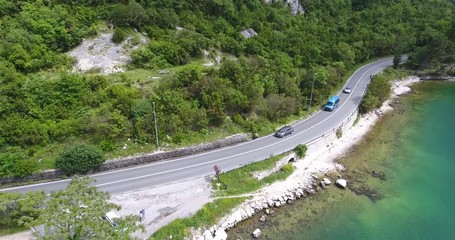 The quadcopter is flying over a narrow asphalt road running along the sea shore. We can see the turquoise sea and the seashore covered with green trees on the other side of the road