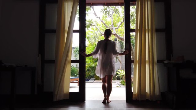 tropical morning. early in the morning, a young woman dressed in a white nightgown of fine material comes to the open door and looks out into the yard. the wind ruffled her nightgown.