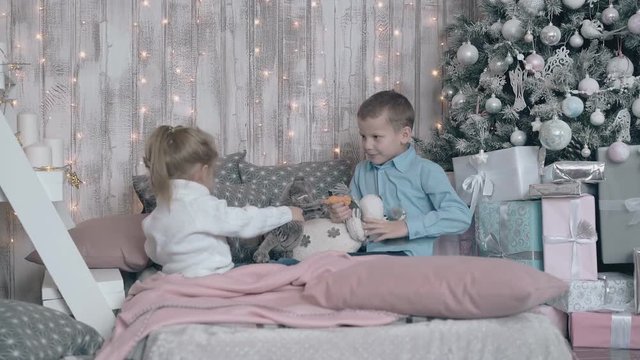 sweet small sister and brother in warm sweaters play with stuffed toys on pink couch near christmas tree and presents