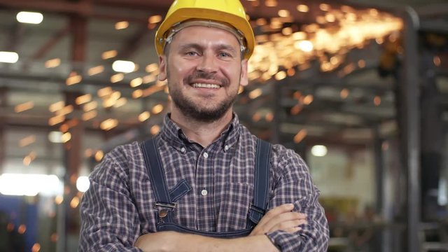 Pan chest-up shot of smiling confident Caucasian engineer, standing in factory workshop with folded arms, wearing plaid shirt, overalls and hardhat, with bright welding sparks falling in background