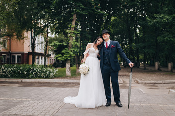 Stylish groom in a blue suit with an umbrella and a hat, and a beautiful bride hugging, on the boulevard in the city. Wedding portrait of newlyweds in love. Concept and photography.