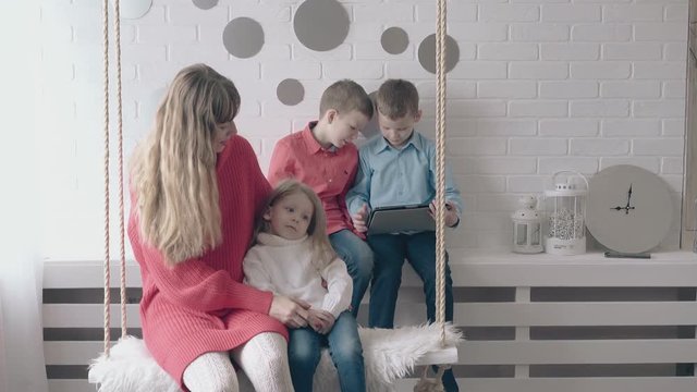 blond mom and daughter ride on white swing near sons playing on tablet against white brick wall by silver clock