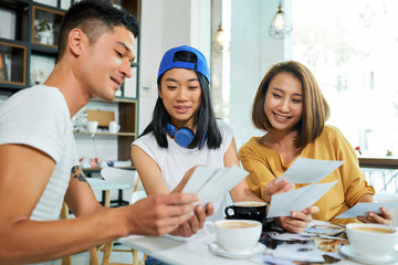 Fototapeta premium Group of positive young Asian people discussing printed photos when drinking coffee in cafe