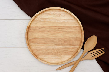 Empty round wooden plate with spoon, fork and dark brown tablecloth on white wooden table. Top view image.