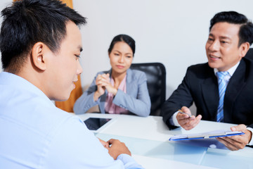Fototapeta premium Mature Vietnamese hr manager pointing at cv and asking questions young applicant at meeting in big company