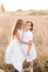 Cute little girls sisters with blond  hair in a summer field at sunset in white dresses with a straw hat