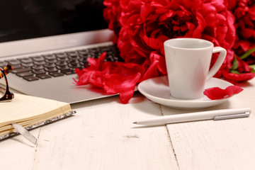 view women's office desk with flowers. Female workspace with laptop, flowers peonies, accessories, notebook, glasses, cup of coffee on white background. Holiday background
