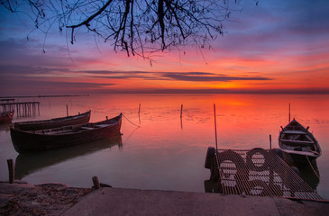 Before the sunrise, red clouds reflecting on the water. Two boats are docked near the shore.