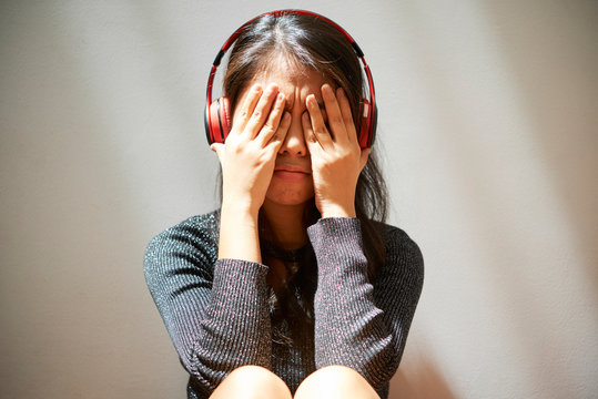Young woman sitting and leaning on the wall in wireless headphones covering her face with hands and crying