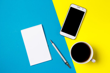 Smartphone, notepad and cup of coffee on a yellow and blue art background. Top view