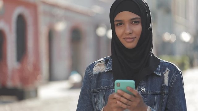 Young muslim woman wearing hijab headscarf stands in the city center uses her phone and looks right to the camera.