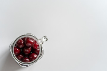 Red juicy cherries in the glass jar on a white table in the left-down corner. Top view