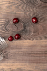 Red juicy cherries scattered from out the glass jar on a brown wooden table from the down-left corner. vertical